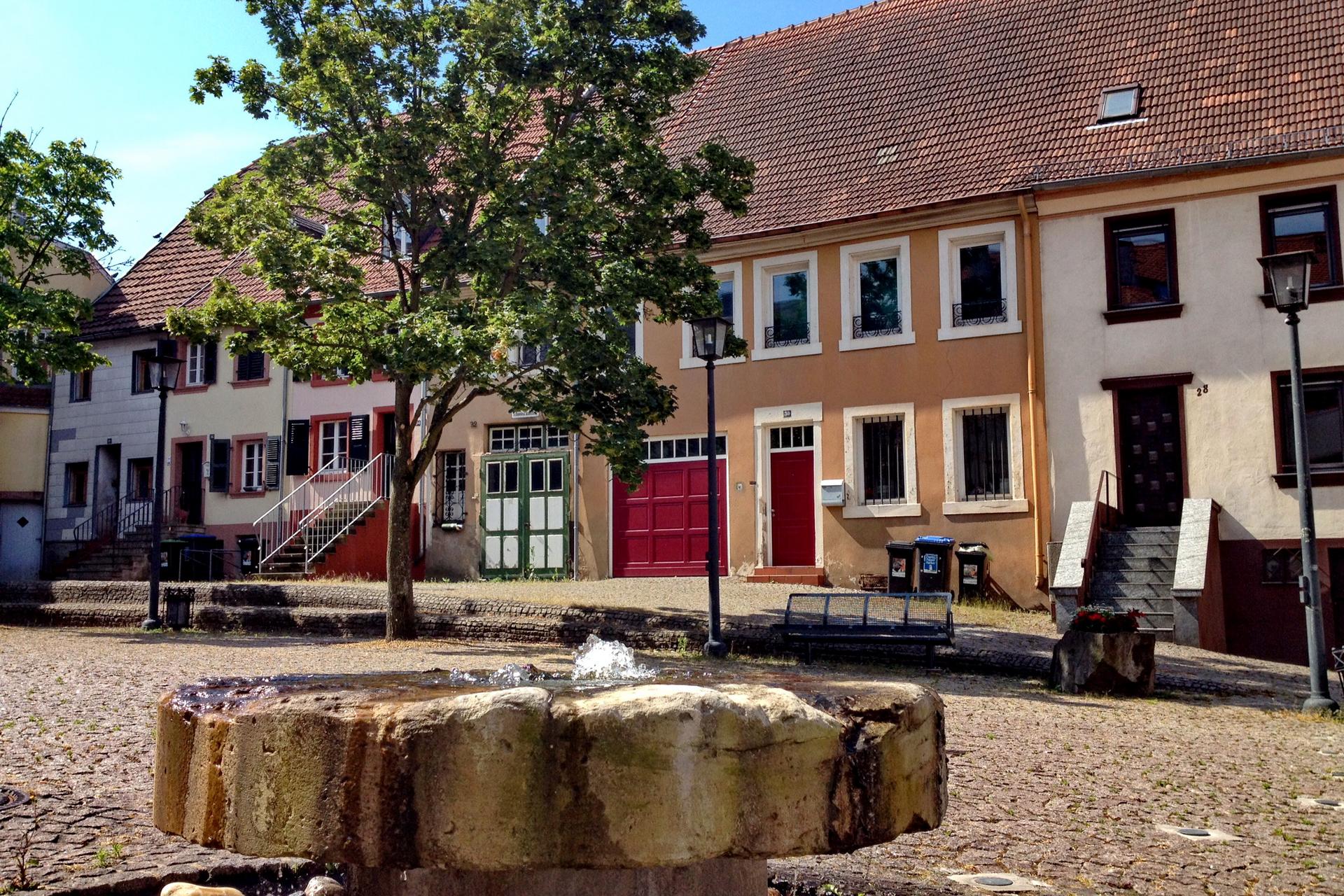 Bunte Handwerkerhäuser im barocken Stil stehen um einen kleinen Platz mit Brunnen in St. Ingbert